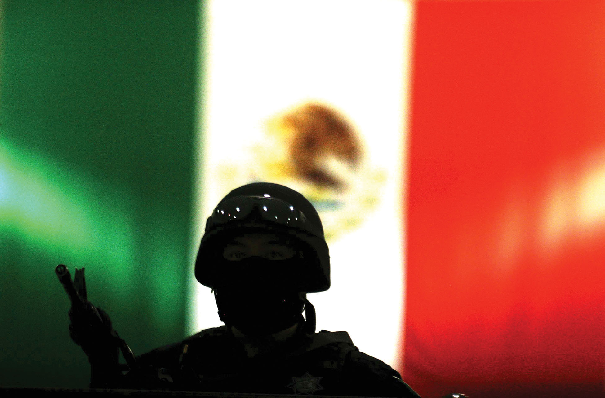 A Mexican federal policeman in tactical gear stands silhouetted before his nation’s flag. (Photo by Jesús Villaseca Pérez.)