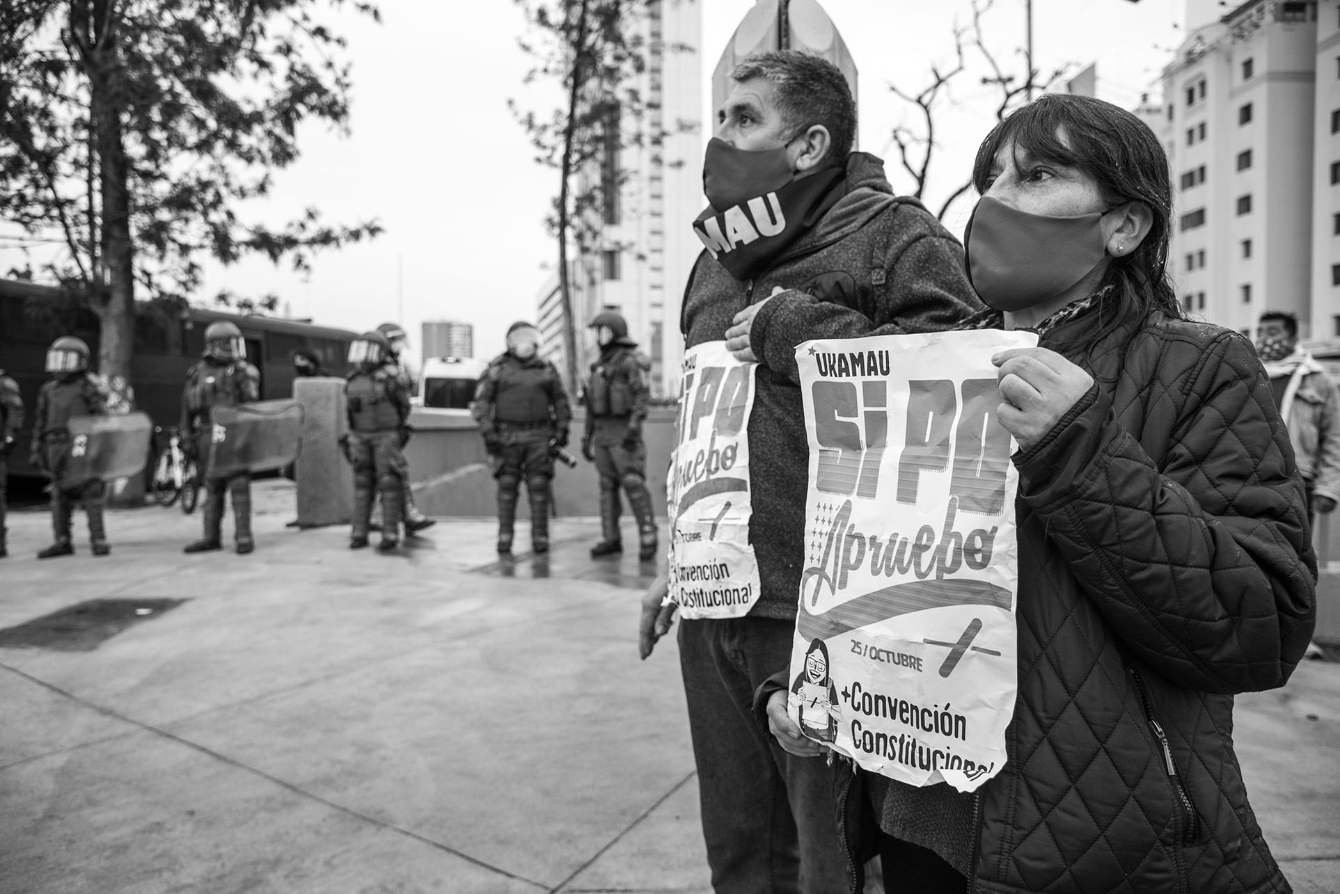 A masked couple supports the campaign for  a new constitution in Chile,  August 2020. (Photo by Paulo Slachevsky.) Standing in front of police in riot gear, a masked mature couple with signs supports the campaign for  a new constitution in Chile,  August 2020. (Photo by Paulo Slachevsky.)