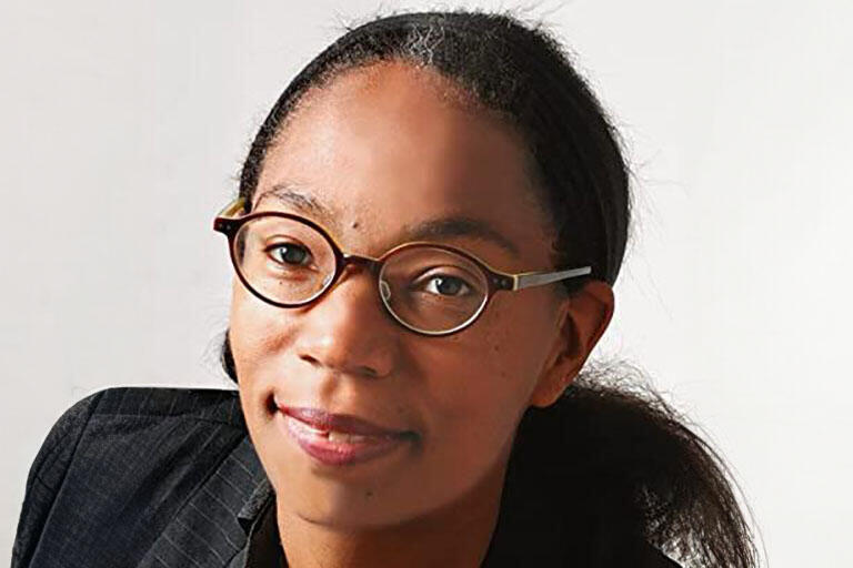 A woman with a black suit, with glasses and black hair in front of a white background