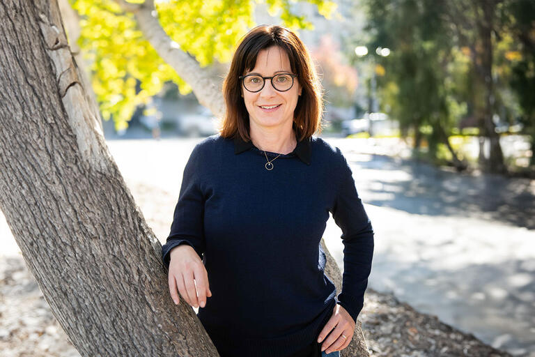 A woman with a dark blue sweater vest with round black glasses. She has Brown hair and is leaning onto a tree facing the camera, with the sun out