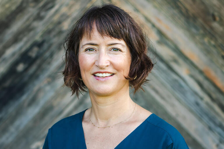 A woman with short brown hair and blue eyes, looking into the camera with a gray wooden blurred background