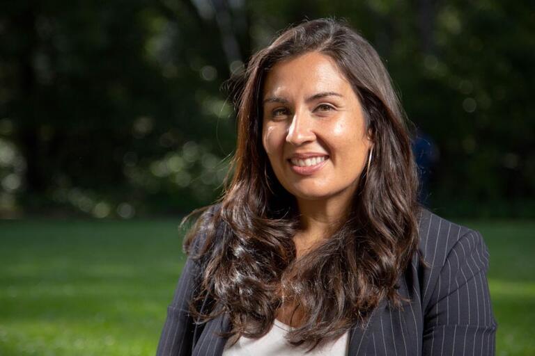 A woman with a suit and long brown hair with hoop earrings, in front of a field