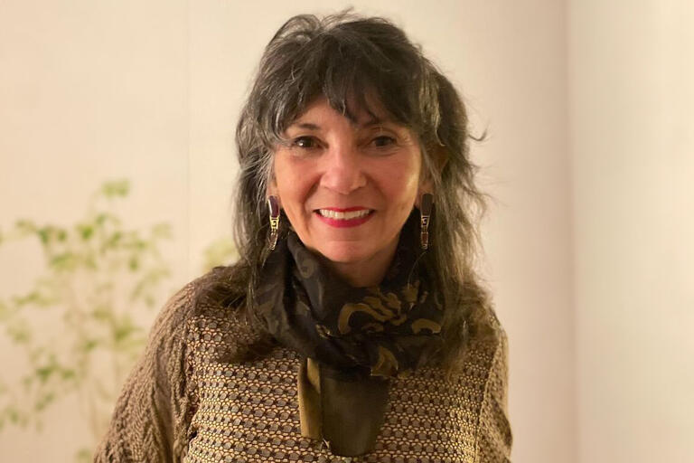 Older woman with a patterned shirt, with a patterned scarf, and gray hair. She is wearing earrings with red lipstick