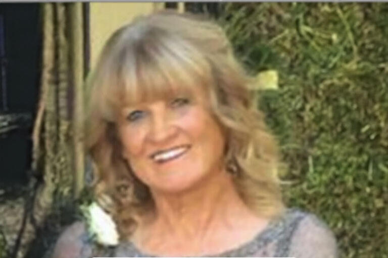 Older woman with blonde hair smiling, in front of some greenery