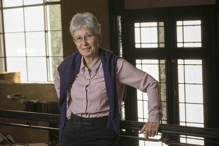 Older woman with a purple vest and a pink button up with black pants. She has glasses, grey hair, and is wearing a necklace