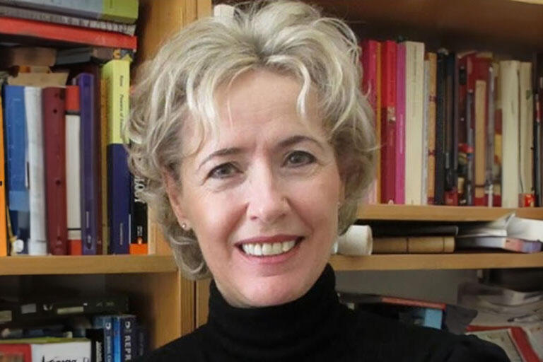 A middle-aged woman with blonde hair, in a black turtleneck. In front of a bookshelf