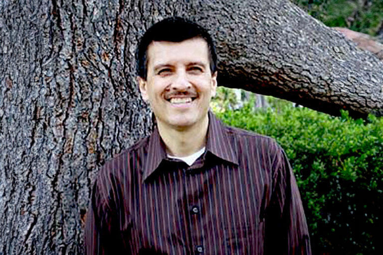 A man with vertical stripped red button up with black hair in front of a tree