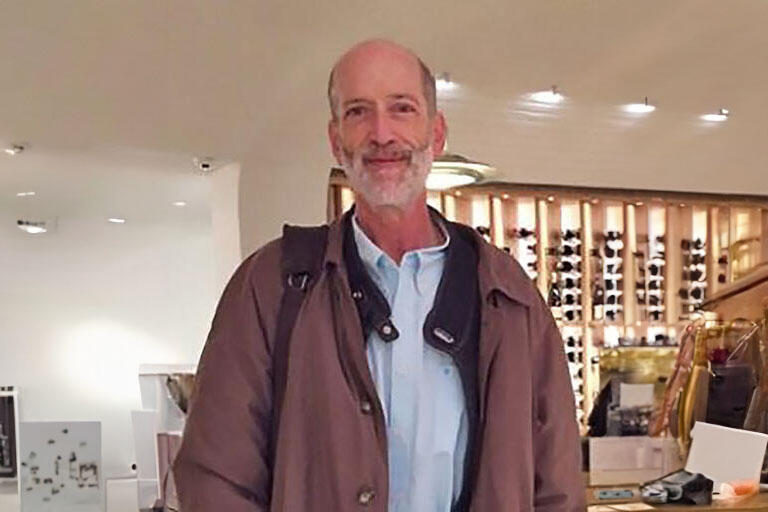 Older man inside a winery, wearing two layers of coats and a blue button up with a white beard