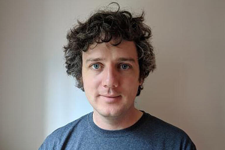 a man with grey-black hair with blue eyes and a grey shirt in front of a gray wall.