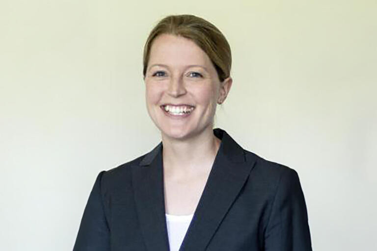 A woman with a black suit, with blonde hair in front of a grey-yellow wall