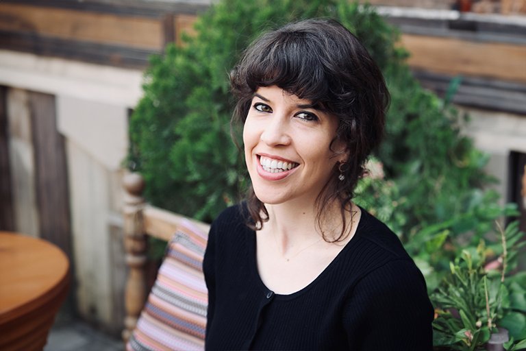 A woman with black hair wearing a black cardigan sitting and in front of some greenery