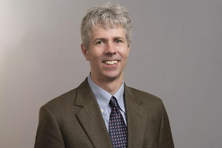 A man in a blue suit, brown jacket over, with grey hair and green eyes