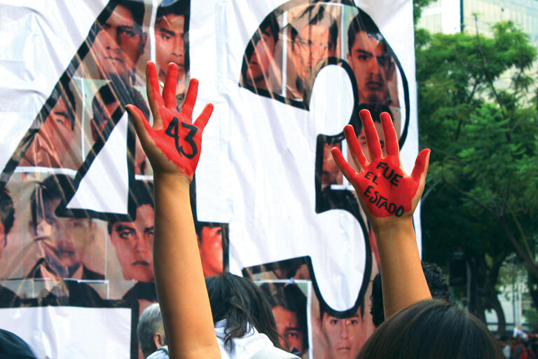 A photo of hands covered in fake blood in front of a poster reading “It was the state” on the third anniversary of the disappearance of 43 students in Iguala, Mexico. (Photo by Adrián Martínez.)