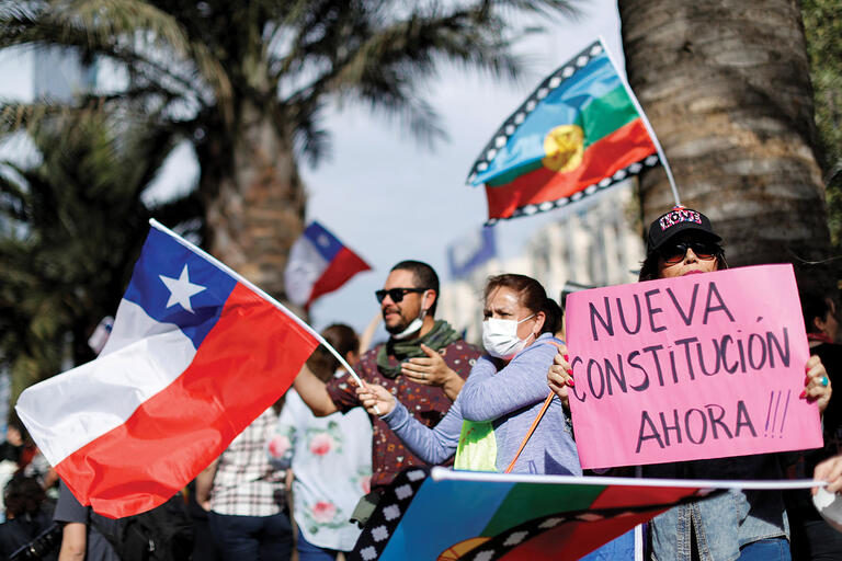 A woman’s sign demands a “New Constitution Now!!!” as Chilean and Mapuche flags fly at a demonstration in Chile, November 2019.  (Photo by Jorge Silva/Reuters.)