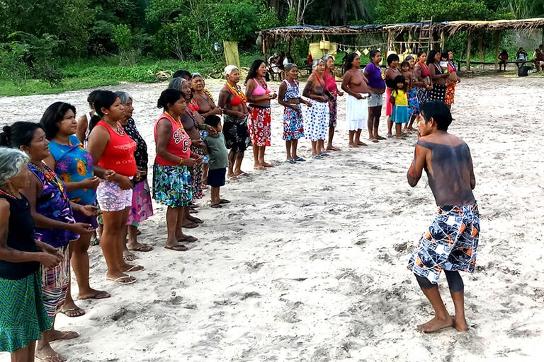 A group of Indigenous women sing and listen to a man play the flute, linked to the article on Panhi musicality.