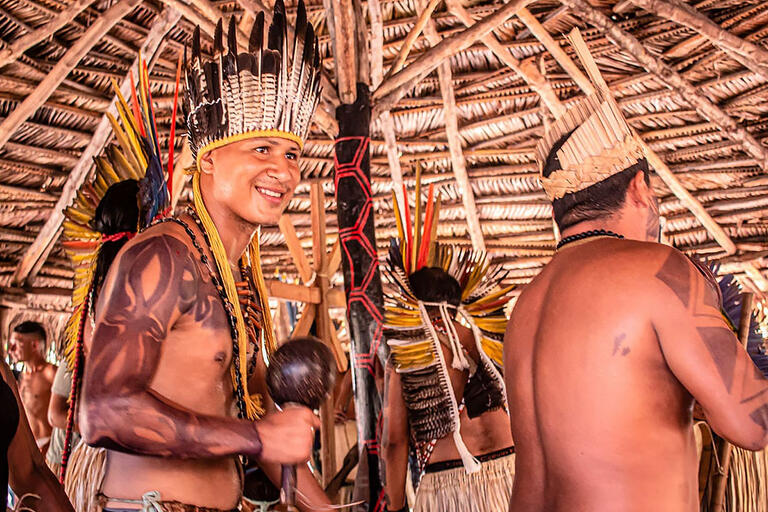 Indigenous people dance in a ceremony, wearing Indigenous headgear and body tattoos, carrying rattles.