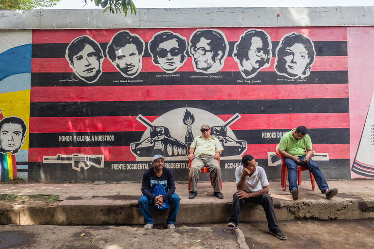 Men sit in front of a black and red Sandinista mural.