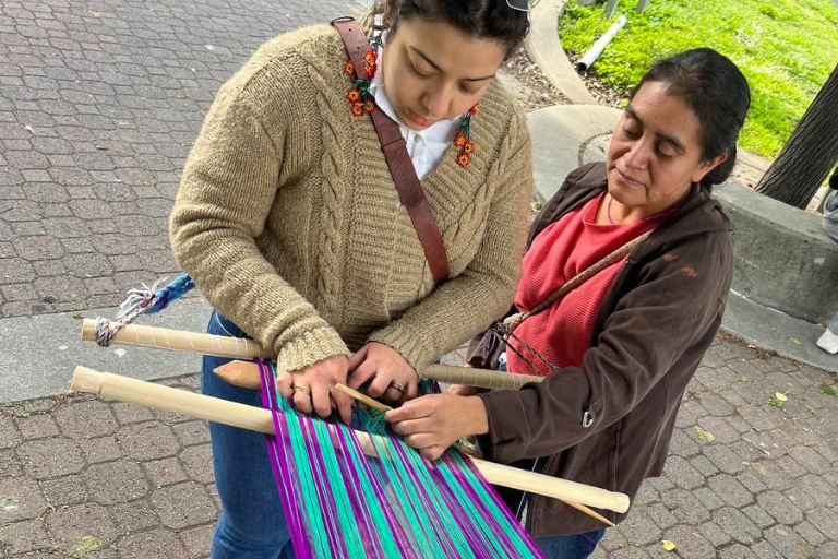 A weaving teacher helps a student weave turquoise and purple yarn on a loom