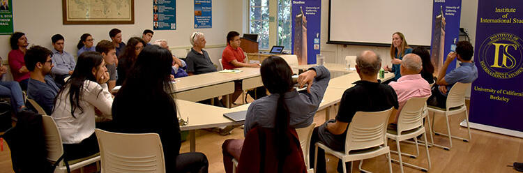 The speaker is sitting in the corner head of a large table, sitting with an  audience in meeting room, while presenting.