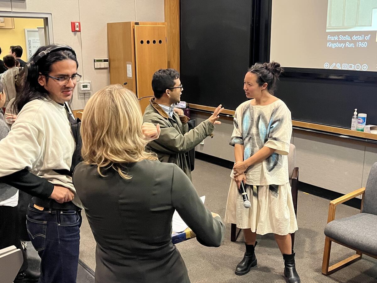 Students chatting after conversation. One students speaks to Irene Small at the front of the classroom.