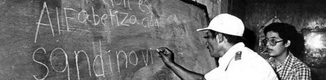 A black and white photo showing a man writing 'Revolucion es Alfebetizacion Sandinista' on a chalkboard, with a woman observing