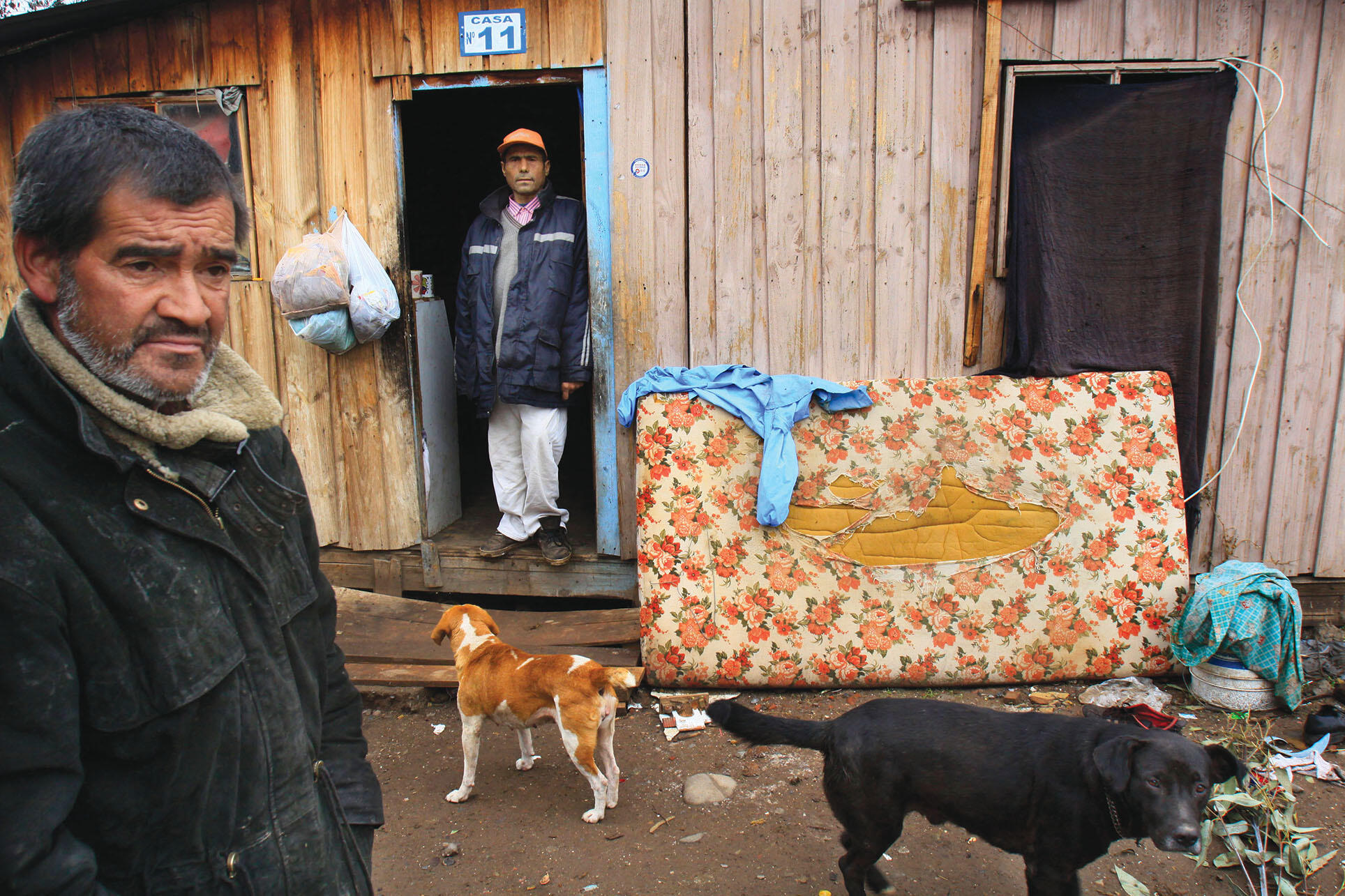 Two men and dogs outside a shack in El Peñon, a shantytown in the Puente Alto sector of Santiago, is home to those left behind. (Photo by Victor Ruiz Caballero/Associated Press.)