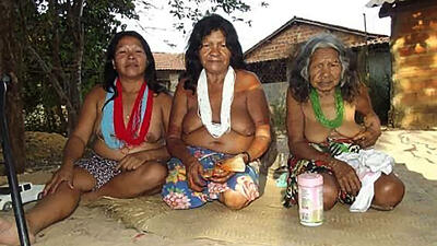 Three women wearing skirts and floral necklaces sit on the ground in front of wooden buildings.