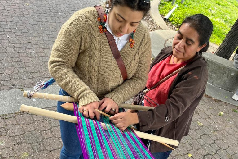 Mam Weaving Class A weaving teacher helps a student weave turquoise and purple yarn on a loom